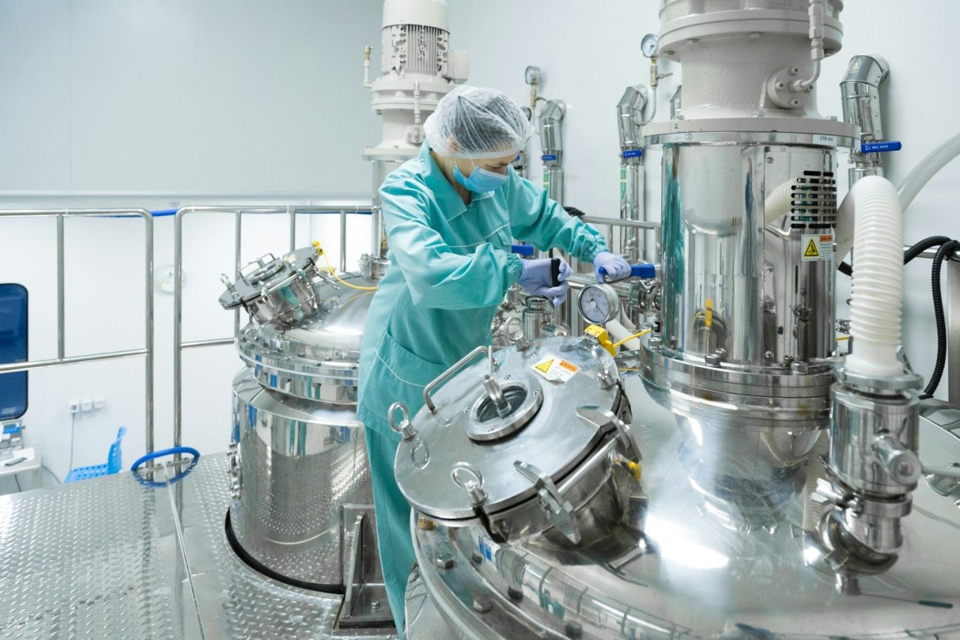 A technician in protective gear works with large stainless steel equipment in a cleanroom.